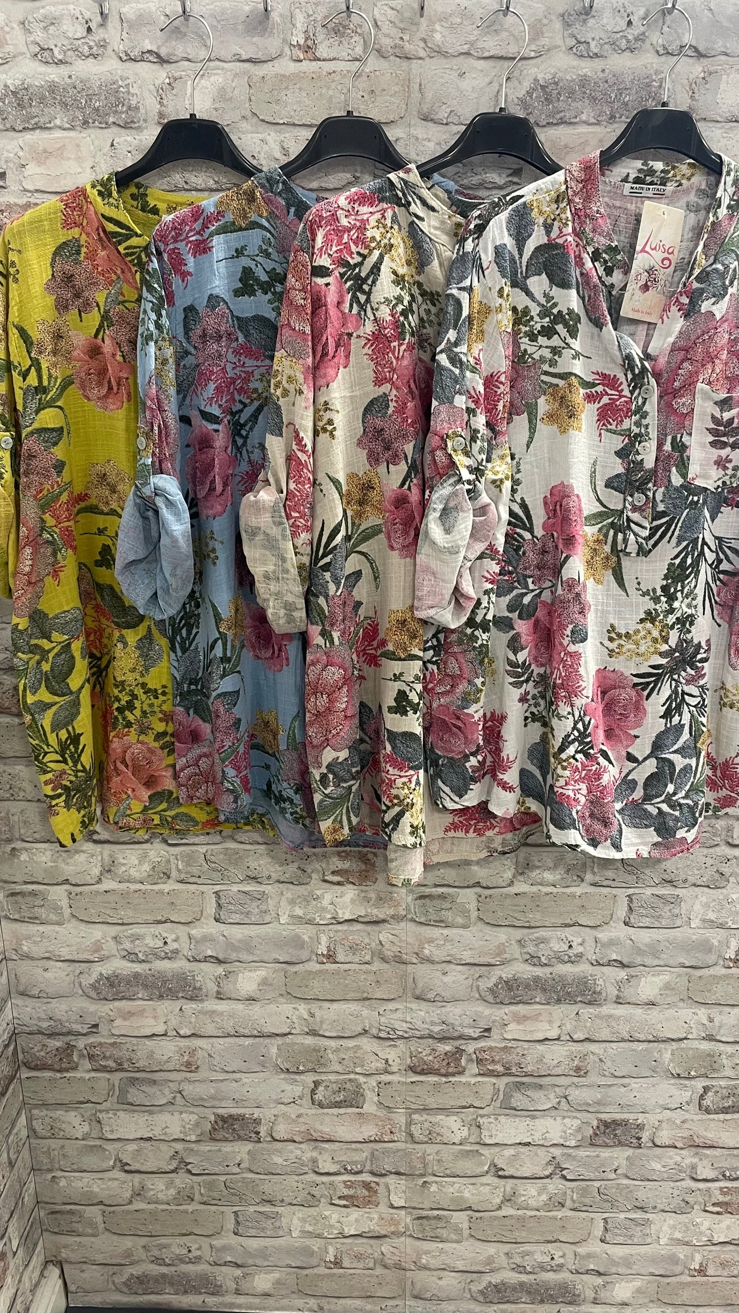English Garden Grandad Shirt in floral patterns, 100% cotton, with single pocket, on hangers against brick wall.