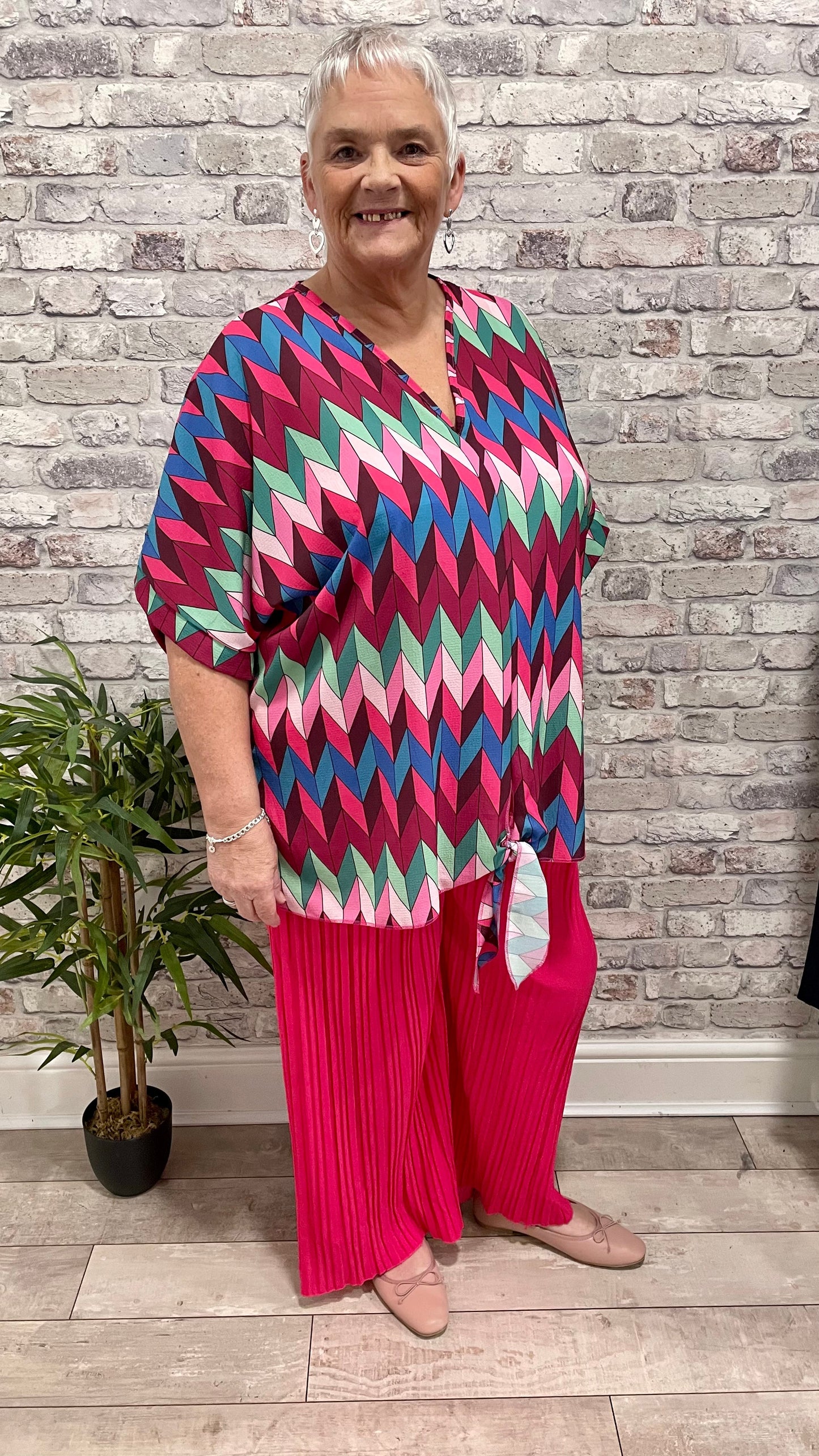 Pleated Palazzo Trouser in vibrant pink worn by a smiling woman, styled with a colorful top, against a brick background.